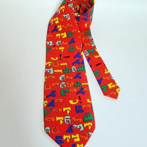 May include: A red tie featuring a repeating pattern of colourful abstract shapes in yellow, blue, green, and orange. The tie is made of a smooth fabric and is displayed against a white background. A classic accessory.