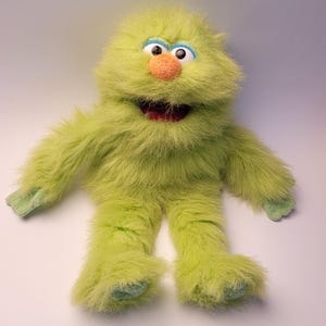 Puede incluir: Un títere de monstruo de peluche color verde lima con pelaje largo y desgreñado, una nariz naranja redonda, ojos grandes blancos con pupilas negras, párpados azules y una boca abierta que muestra un interior oscuro. Brazos y piernas largos.