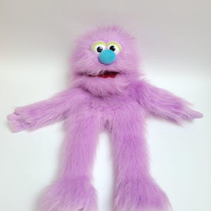 May include: A plush puppet covered in long, shaggy lavender fur. It features two large white eyes with black pupils, a bright blue rounded nose, and a wide mouth with a red interior. The puppet has two long arms and two long legs.