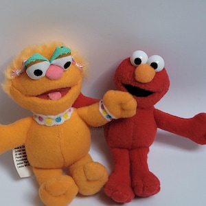 May include: Two plush Sesame Street character toys. One is orange with a pink nose, a white and colorful collar, and a green and white headband. The other is red with a large orange nose and white eyes.
