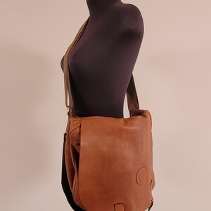 May include: A tan leather messenger bag with a large front flap, a long adjustable shoulder strap, and a circular embossed logo on the lower right corner. The bag features a single vertical leather strap closure hanging from the center front.