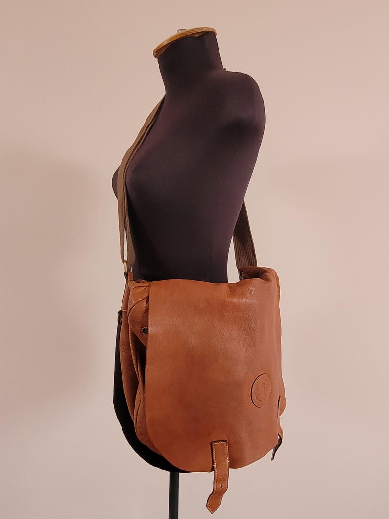 May include: A tan leather messenger bag with a large front flap, a long adjustable shoulder strap, and a circular embossed logo on the lower right corner. The bag features a single vertical leather strap closure hanging from the center front.