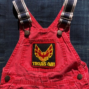 May include: Red overalls with adjustable straps and a black and yellow embroidered patch that says "TRANS AM" with a bird design.