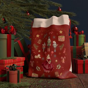 Puede incluir: Bolsa de regalo roja de Navidad con cordón de ajuste blanco. La bolsa está decorada con un tema de Cascanueces y presenta ilustraciones de un cascanueces, una bailarina, bastones de caramelo y otros elementos navideños.