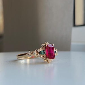 May include: A gold ring with a large, rectangular, red gemstone surrounded by smaller, clear and green gemstones. The ring has a delicate, floral design.