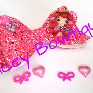 May include: Pink glitter hair bow featuring a small doll figurine wearing a pink dress and a floral crown. The bow is decorated with pink flowers and a rhinestone. The image also includes three pink heart-shaped embellishments and three pink bows.