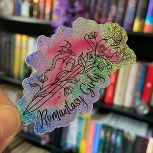 Puede incluir: Una pegatina holográfica con un fondo degradado rosa y azul. La pegatina presenta un dibujo de línea negra de una daga con un dragón envuelto alrededor. La daga está rodeada de flores y el texto "Romantasy Girl" está escrito en una fuente cursiva.