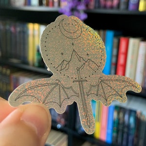 May include: A silver glitter sticker with a black line drawing of a sword, a mountain, a crescent moon, and wings. The sticker has a holographic finish.