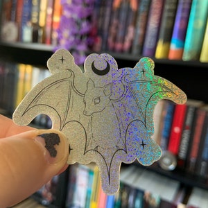 May include: A silver holographic sticker with a black outline of a bat with wings spread wide. The bat is facing forward with a crescent moon above it. There are two small stars on either side of the bat.