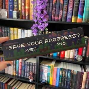 Può includere: Un segnalibro in stile gioco pixelato con sfondo blu e stelle bianche. Il segnalibro recita "Save Your Progress?" con le opzioni "Yes" e "No" in testo bianco. Il segnalibro è tenuto in una mano contro una libreria con molti libri.