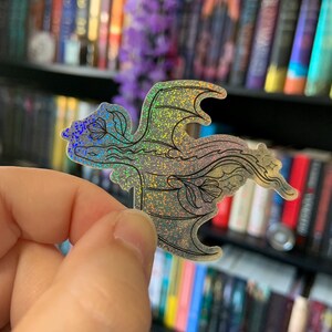 Könnte beinhalten: Ein holografischer Aufkleber in Form eines Drachen mit floralen Akzenten. Der Aufkleber hat ein schimmerndes, irisierendes Finish mit schwarzen Umrissen. Der Drache wird vor einem verschwommenen Hintergrund aus Büchern und lila Blumen gehalten.