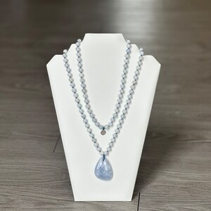 May include: A light blue gemstone necklace with a teardrop pendant on a white display stand. The necklace is made of multiple strands of beads.