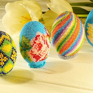 May include: Four beaded Easter eggs with different designs. The eggs are blue, yellow, green, and pink with white accents. The eggs are arranged in a row on a white wooden surface.