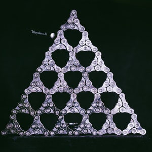 Może przedstawiać: Trójkątny wzór wykonany ze srebrnych łańcuchów rowerowych na ciemnym tle. Łańcuchy są ułożone w kształt geometryczny. Widoczny jest tekst "tricycleco.fr".