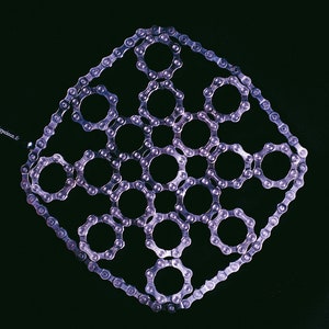 Może przedstawiać: Geometryczny wzór w kształcie rombu wykonany z łańcuchów rowerowych, z okrągłymi wzorami wewnątrz. Łańcuchy są srebrne na ciemnym tle. Tekst "Tricyclea 6" jest widoczny w rogu.