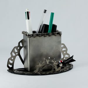 May include: A metal desk organizer with a chain design around the top of the container. The organizer is made from recycled bicycle parts and has a black tag with the text "Tricyleco" on it.
