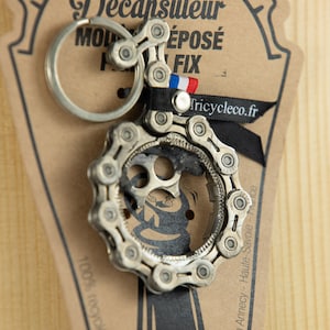 Peut inclure: Un ouvre-bouteille en forme de chaîne de vélo en argent. Le porte-clés est attaché à une carte brune avec le texte "Decapsuleur Molepose Fix" et "Tricycleco.fr".