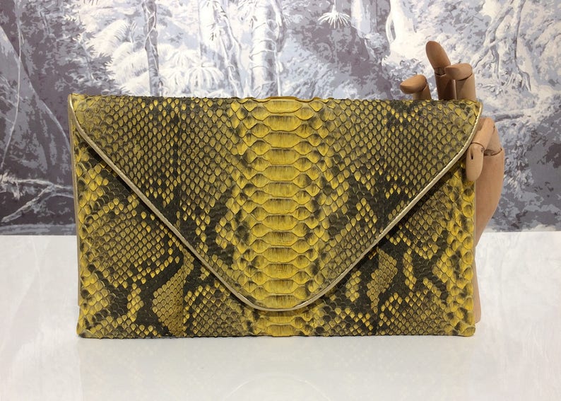 K&ouml;nnte beinhalten: Eine gelbe und schwarze Schlangenleder-Clutch mit goldfarbenem Verschluss.