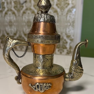 May include: A small, ornate copper and brass teapot with intricate detailing. The teapot features a pointed lid, a curved spout, and a handle with a chain. The body of the teapot has engraved patterns and a decorative base.
