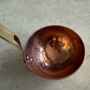 May include: A copper ladle with a hammered bowl and a brass handle. The bowl has a reflective surface, and the handle is attached with rivets. The ladle is designed for kitchen use.