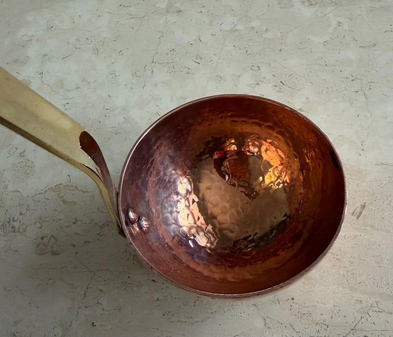 May include: A copper ladle with a hammered bowl and a brass handle. The bowl has a reflective surface, and the handle is attached with rivets. The ladle is designed for kitchen use.