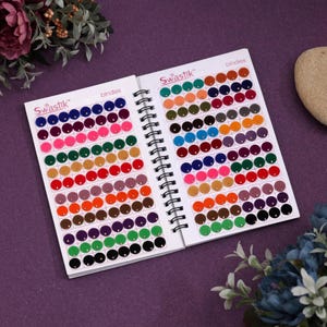 May include: Two open spiral-bound swatch books with a variety of colorful, round stickers. The stickers are arranged in rows and columns, displaying a spectrum of colors including pink, orange, green, and purple. The books are labeled "Swastik" and "binders".