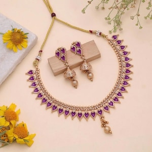May include: A gold-toned necklace and matching pair of drop earrings featuring purple, teardrop-shaped accents. The necklace has a delicate chain with a central pendant drop, while the earrings consist of a stud top and a bell-shaped base.
