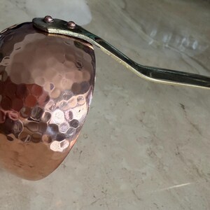 May include: A copper ladle with a hammered texture on the bowl. The handle is a silver color and is attached to the bowl with rivets. The ladle is designed for kitchen use.