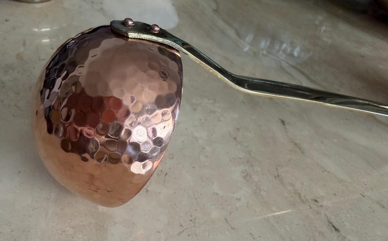 May include: A copper ladle with a hammered texture on the bowl. The handle is a silver color and is attached to the bowl with rivets. The ladle is designed for kitchen use.
