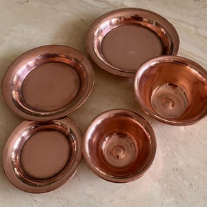 May include: A set of five polished copper vessels consisting of three shallow circular plates and two deep rounded bowls. Each piece features a smooth, reflective metallic surface with a warm reddish-brown tone and a simple, clean rim design.