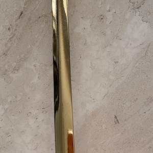 May include: A copper and brass ladle with a hammered bowl and a long, shiny handle. The handle is gold-colored and has a slight curve. The ladle is likely used for cooking or serving.