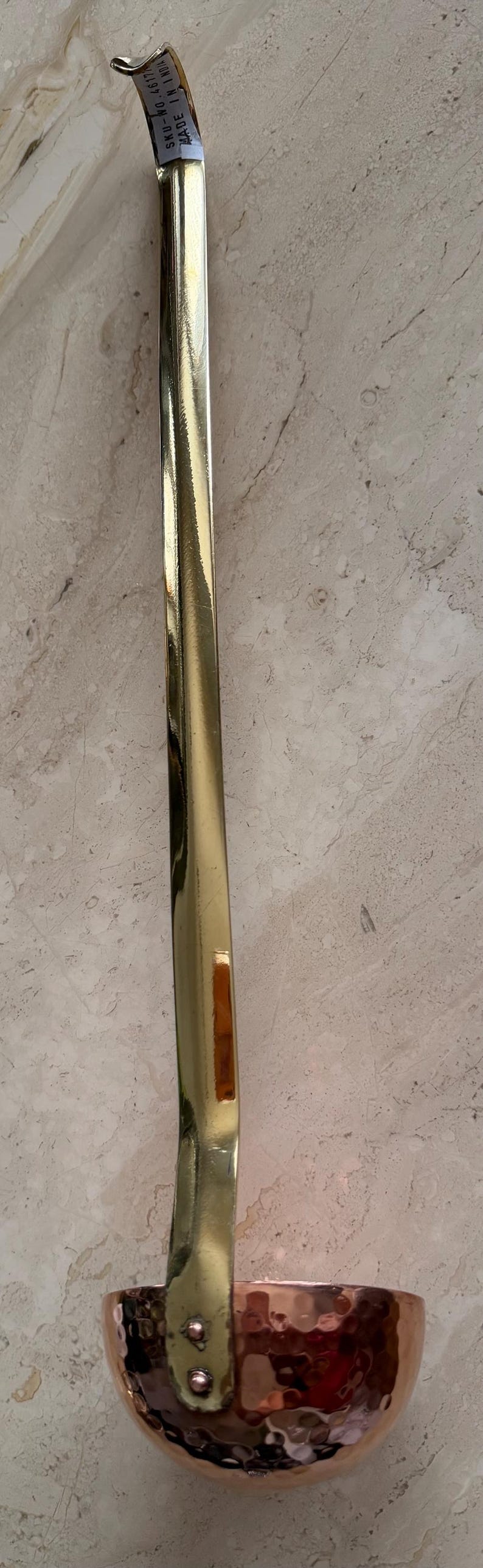 May include: A copper and brass ladle with a hammered bowl and a long, shiny handle. The handle is gold-colored and has a slight curve. The ladle is likely used for cooking or serving.