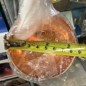 May include: A copper-colored, round bowl is partially wrapped in clear plastic. A yellow measuring tape shows the bowl's diameter is approximately 7 inches. The bowl is likely made of metal and has a reflective surface.