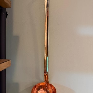 May include: A copper ladle with a long handle and a hammered bowl. The ladle is hanging on a light green wall. The handle has a hook at the end for hanging.