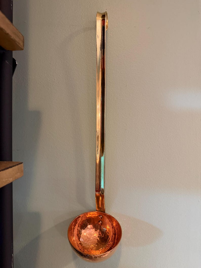 May include: A copper ladle with a long handle and a hammered bowl. The ladle is hanging on a light green wall. The handle has a hook at the end for hanging.
