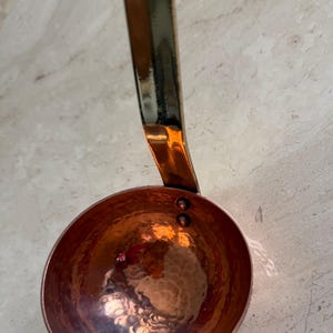 May include: Copper ladle with a hammered bowl and a long handle. The ladle has a shiny, reflective surface and a warm, reddish-brown color. The handle is a darker, metallic color.