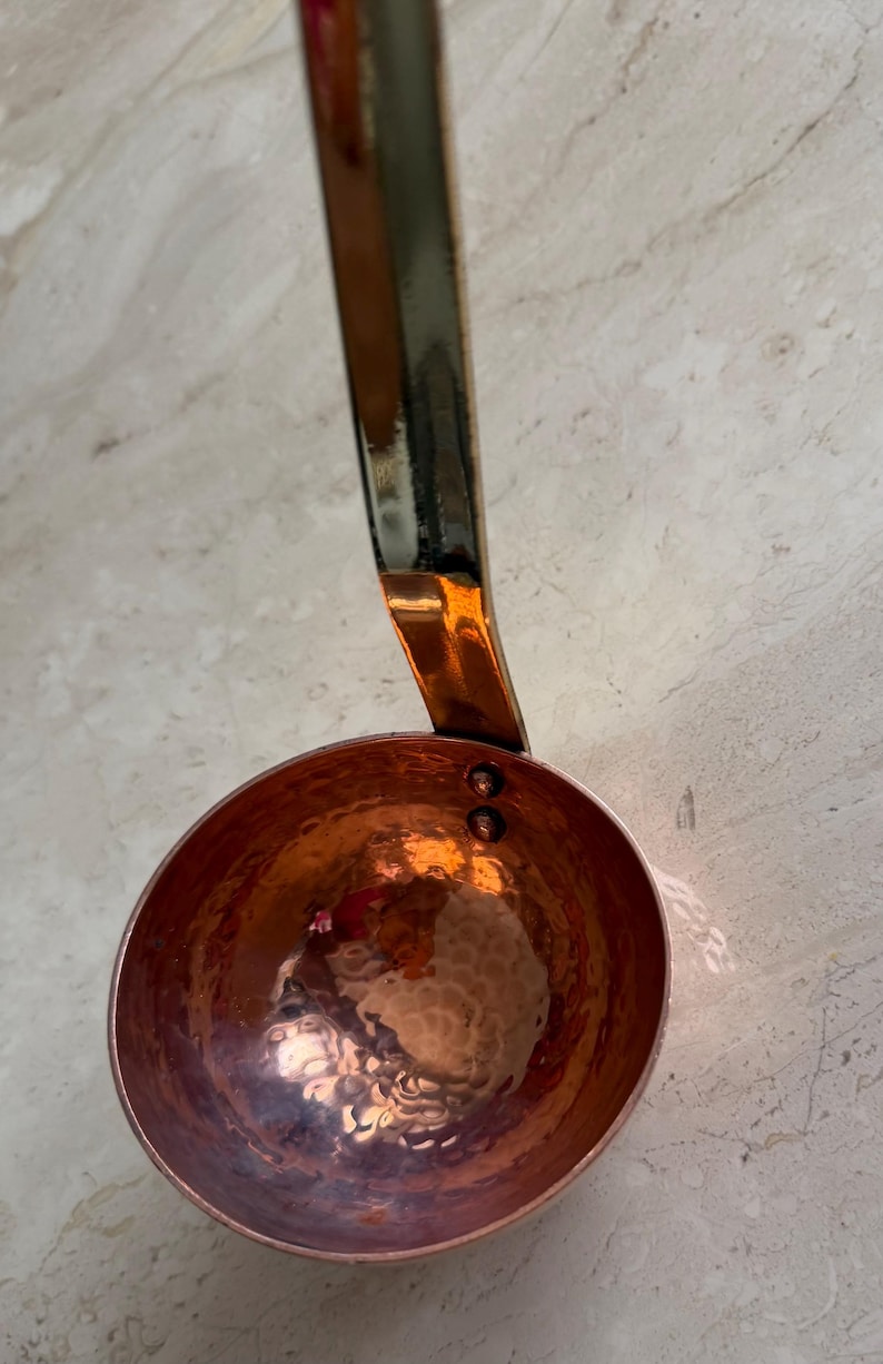 May include: Copper ladle with a hammered bowl and a long handle. The ladle has a shiny, reflective surface and a warm, reddish-brown color. The handle is a darker, metallic color.