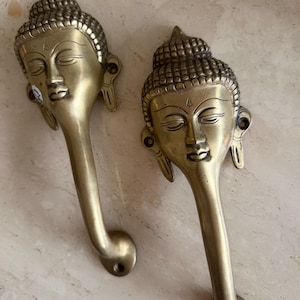 May include: Two brass door handles, each featuring a detailed Buddha head design. The handles have a curved shape and a warm, golden hue, adding an elegant touch to any door. The Buddha's serene expression is captured in the intricate details.