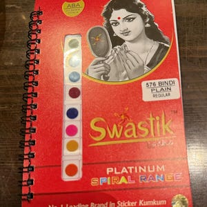 May include: A red spiral-bound booklet containing a vertical strip of colorful circular stickers. The cover features an illustration of a person holding a mirror, the text Swastik, 576 BINDI PLAIN REGULAR, PLATINUM SPIRAL RANGE, and No. 1 Leading Brand in Sticker Kumkum.
