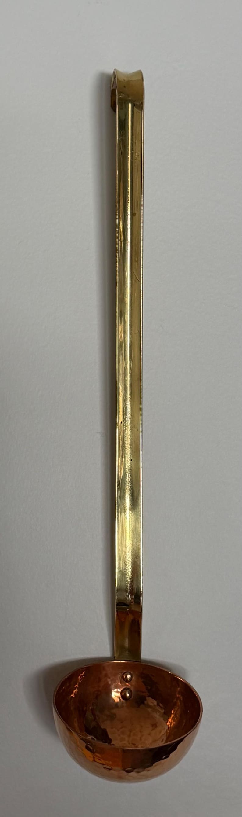 May include: A copper ladle with a brass handle. The ladle's bowl is a warm copper color, while the handle is a shiny brass. The handle is long and slender, with a curved end for hanging. A kitchen utensil.