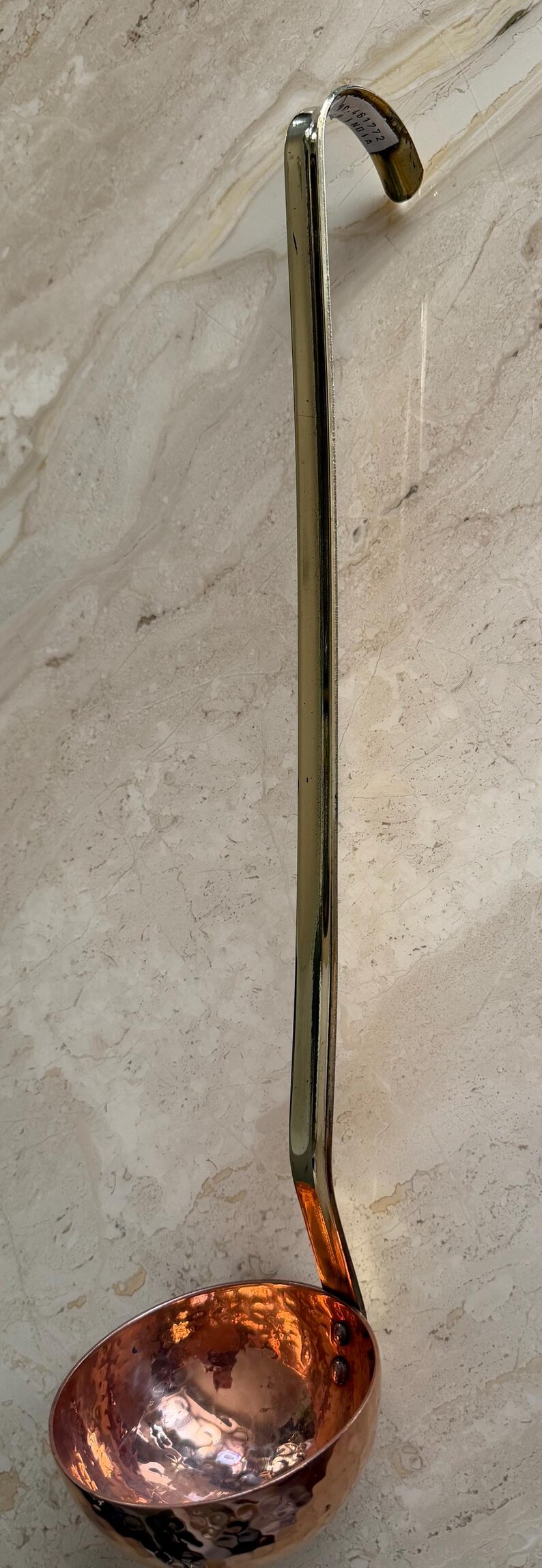 May include: A copper-colored ladle with a long, shiny, silver-colored handle. The ladle's bowl has a hammered texture. The handle curves at the top for hanging. The ladle is designed for kitchen use.