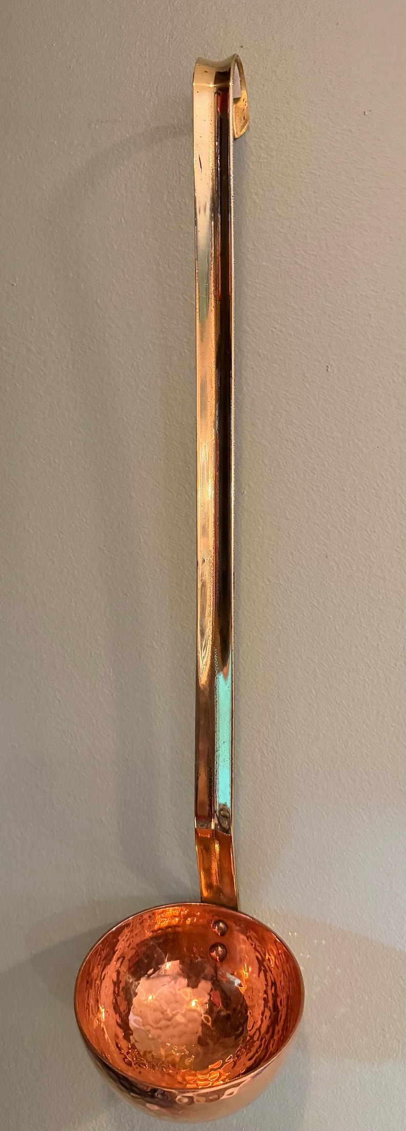 May include: A copper ladle with a hammered bowl and a long, straight handle. The ladle has a shiny, reflective surface and is designed for kitchen use. The handle has a curved hook for hanging.