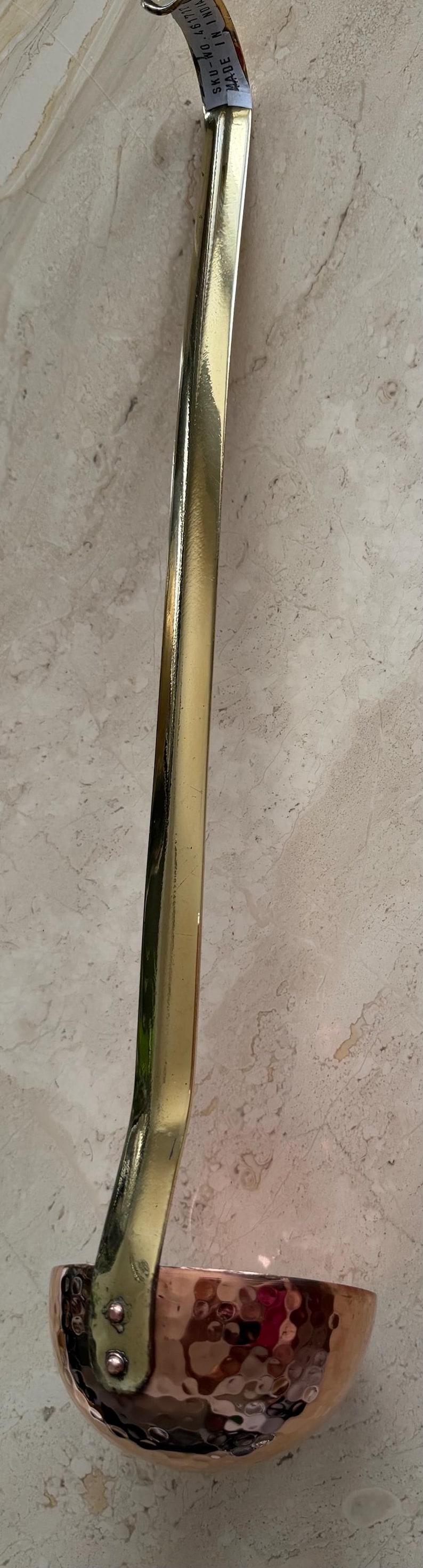 May include: A copper and brass ladle with a long, curved handle. The handle is gold-colored, and the bowl is a shiny copper. The ladle is designed for kitchen use and is likely used for serving soups or sauces.