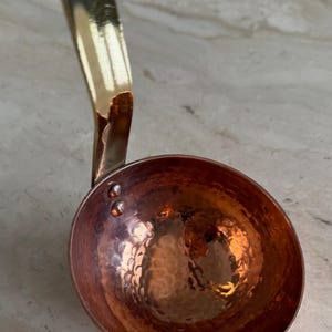 May include: A copper ladle with a hammered bowl and a brass handle. The ladle has a shiny, reflective surface and is designed for kitchen use. The handle curves upward, and the bowl is round with a textured surface.