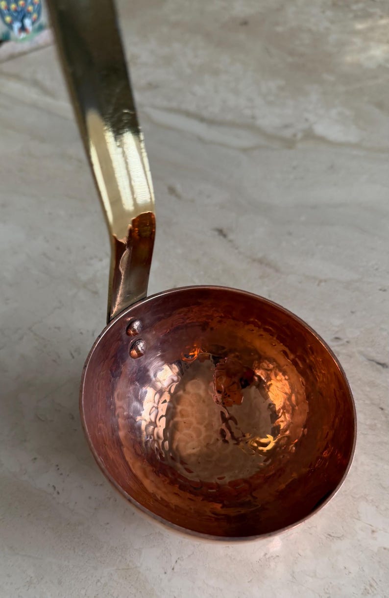 May include: A copper ladle with a hammered bowl and a brass handle. The ladle has a shiny, reflective surface and is designed for kitchen use. The handle curves upward, and the bowl is round with a textured surface.