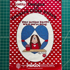 May include: A red booklet with a white heart pattern titled Cathy Designs in Counted Cross Stitch. The cover features a cartoon illustration of a character with long brown hair, a red shirt, and a potted plant, with the text Your mother knows what you're doing. Published by Paragon Needlecraft in 1983.