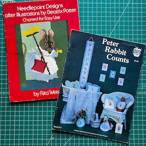 May include: Two softcover needlepoint pattern books. The red book is titled Needlepoint Designs after Illustrations by Beatrix Potter Charted for Easy Use by Rita Weiss. The dark blue book is titled Peter Rabbit Counts by Green Apple Co.