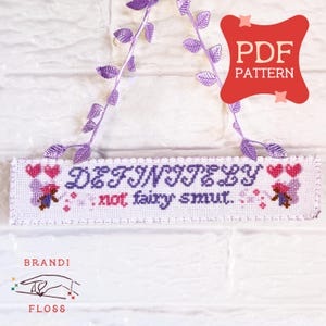 May include: A white cross-stitch sign with the words "DEFINITELY not fairy smut" in purple and red. The sign has a scalloped edge and is decorated with hearts and floral accents. A purple leaf garland hangs from the top.