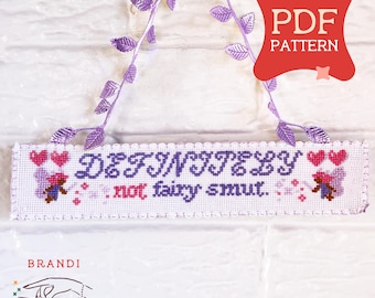以下が含まれることがあります： 紫と赤で「DEFINITELY not fairy smut」という言葉が書かれた白いクロスステッチのサイン。 サインはスカラップエッジで、ハートと花のアクセントで飾られています。 紫色の葉のガーランドが上から吊り下げられています。