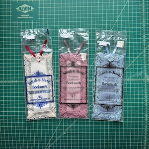 May include: Three Stitch-n-Mark Designer Collection bookmarks in individual clear plastic sleeves. Each bookmark features Aida fabric with lace edging, a ribbon at the top, and printed text. Colors include white with red ribbon, pink with pink ribbon, and blue with blue ribbon.